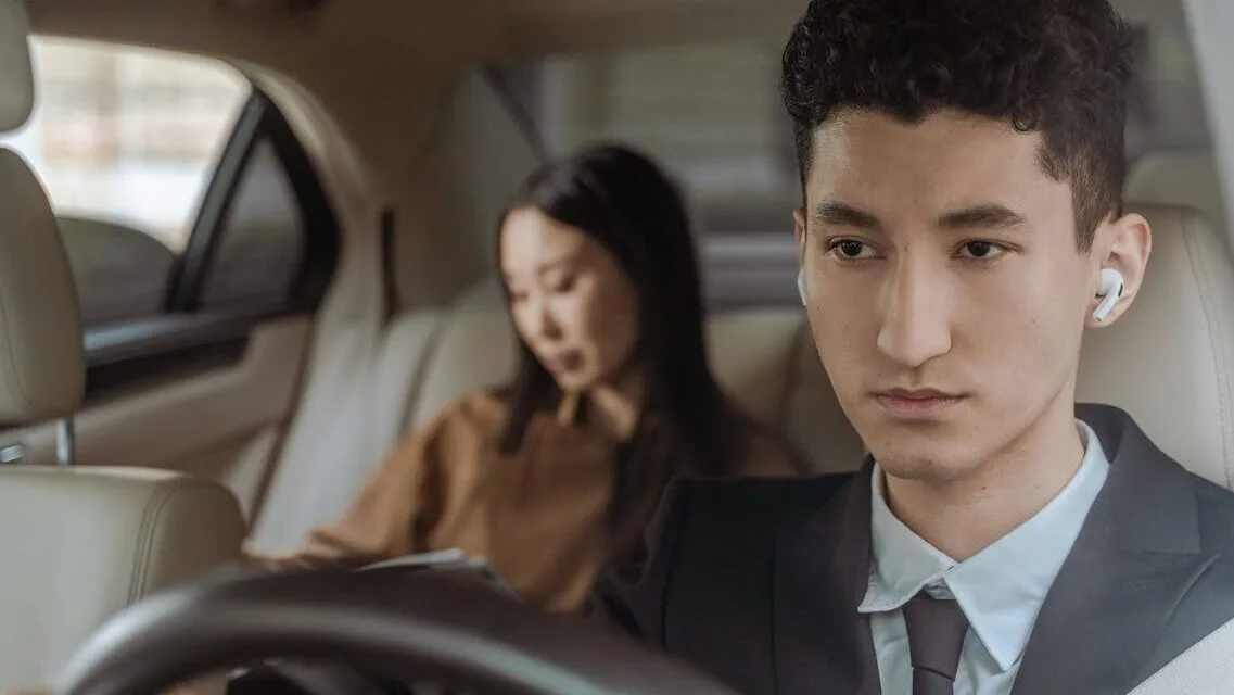 Young professional driver wearing suit and earpiece in luxury car interior with female passenger.